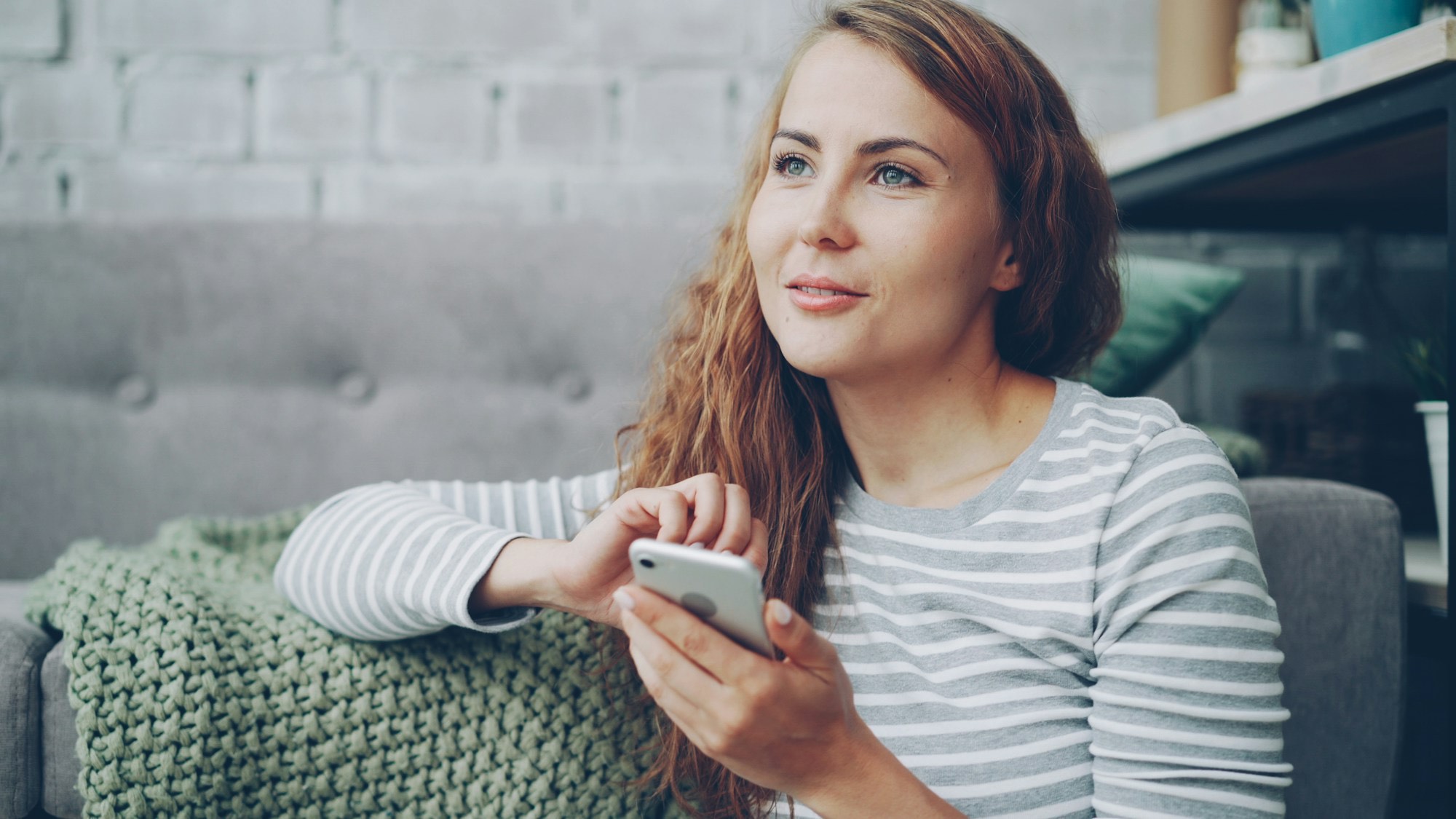 A woman on a couch holding her phone, gazing away in thought